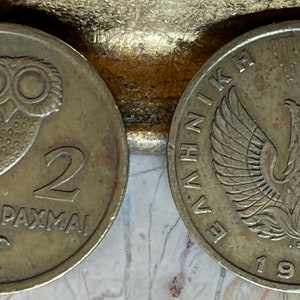 May include: Two vintage Greek drachma coins. One coin features an owl and the number 2, with the word "DRACHMAI" below. The other coin shows a phoenix and the year 1973, with Greek text around the edge. Both coins are silver in color.