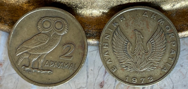 May include: Two vintage Greek drachma coins. One coin features an owl and the number 2, with the word "DRACHMAI" below. The other coin shows a phoenix and the year 1973, with Greek text around the edge. Both coins are silver in color.