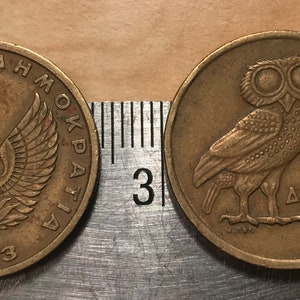 May include: Two bronze Greek coins. One coin features a phoenix with outstretched wings and the year 1973. The other coin shows an owl and the number 2, with Greek text. The coins are next to a ruler.