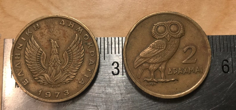May include: Two bronze Greek coins. One coin features a phoenix with outstretched wings and the year 1973. The other coin shows an owl and the number 2, with Greek text. The coins are next to a ruler.