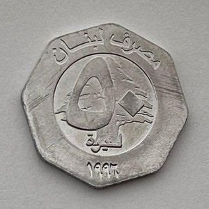 May include: A silver coin with the year 1997 and Arabic text on it. The coin features a stylized image of a cedar tree, a symbol of Lebanon.