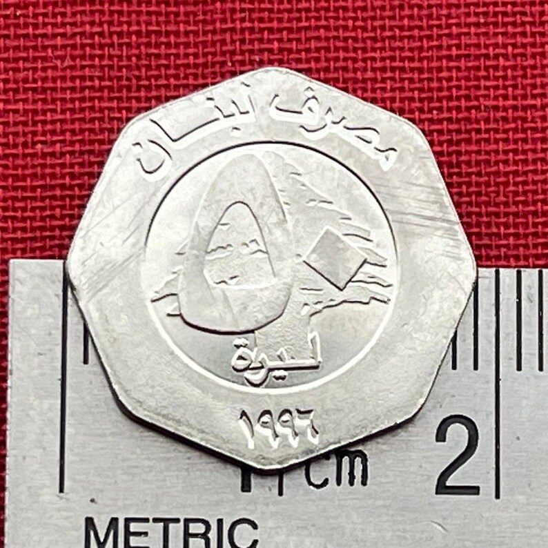 May include: A silver-colored coin with the text "لبنان" and "1997" on it. The coin features a stylized image of a cedar tree and the Arabic word "لبنان" in the center.