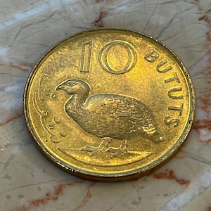 Può includere: Una moneta dorata con il numero 10 e la parola "BUTUTS" incisa sul bordo. Un uccello è raffigurato al centro della moneta.