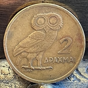 Può includere: Moneta di bronzo raffigurante un gufo con grandi occhi, rivolto verso sinistra. Il numero "2" è a destra, con la parola greca "ΔΡΑΧΜΑ" sotto. La moneta ha forma circolare e un aspetto leggermente usurato.