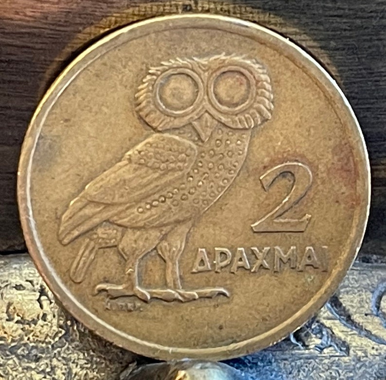 May include: A bronze coin featuring an owl with large eyes, facing left. The number "2" is on the right, with the Greek word "&Delta;&Rho;&Alpha;&Chi;&Mu;&Alpha;" below. The coin has a circular shape and a slightly worn appearance.