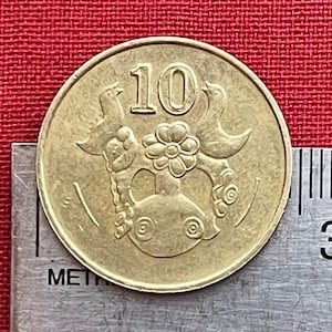 Könnte beinhalten: Eine goldfarbene Münze mit der Zahl "10" in der Mitte und zwei Vögeln auf beiden Seiten der Zahl. Die Vögel schauen sich an und haben ihre Flügel ausgebreitet. In der Mitte der Münze befindet sich eine Blume unterhalb der Zahl.