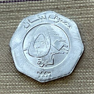 May include: A silver coin with the year 1997 and the Arabic text "جمهورية لبنان" (Republic of Lebanon) on it. The coin features a stylized image of a cedar tree, a symbol of Lebanon.