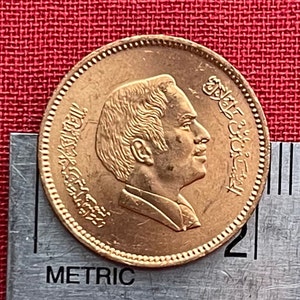 Puede incluir: Una moneda de color dorado con el retrato de un hombre de perfil en el anverso. La moneda tiene texto árabe alrededor del borde.