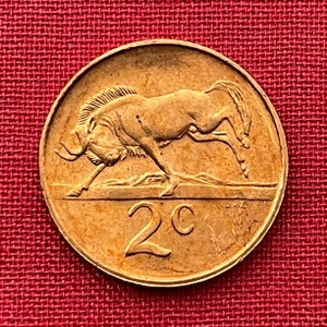 Puede incluir: Una moneda de 2 centavos sudafricana de color dorado que presenta un ñu corriendo hacia la derecha. La moneda tiene la inscripción "2c" en la parte inferior.