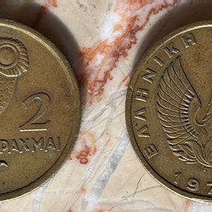 May include: Two vintage Greek coins. One coin features an owl and the number 2 with the word "DRACHMAI". The other coin has a phoenix with the text "ELLINIKI DIMOKRATIA" and the year 1973.