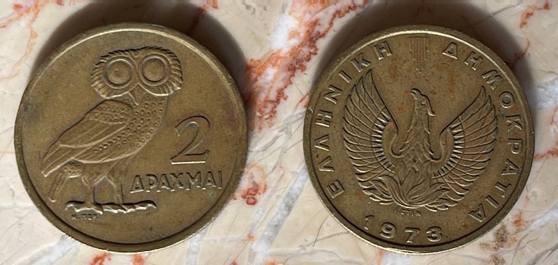 May include: Two vintage Greek coins. One coin features an owl and the number 2 with the word "DRACHMAI". The other coin has a phoenix with the text "ELLINIKI DIMOKRATIA" and the year 1973.