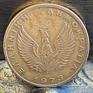 May include: A bronze coin featuring a detailed depiction of a phoenix with outstretched wings, surrounded by Greek text. The year "1973" is prominently displayed below the phoenix. The coin has a worn, antique appearance.