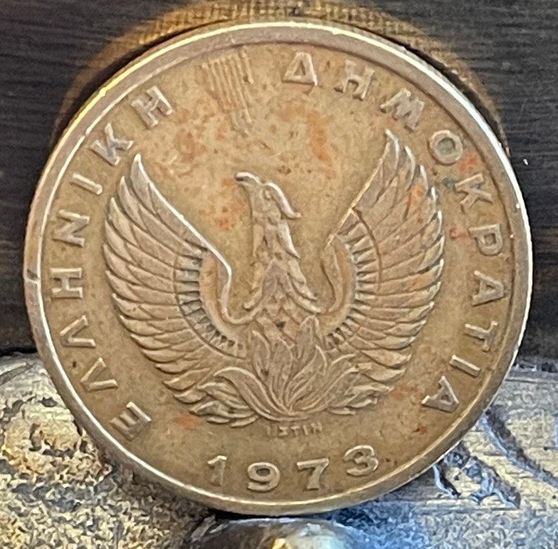 May include: A bronze coin featuring a detailed depiction of a phoenix with outstretched wings, surrounded by Greek text. The year "1973" is prominently displayed below the phoenix. The coin has a worn, antique appearance.