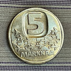 May include: A silver Finnish 5 markkaa coin with the number "5" in the center and the word "MARKKAA" around the edge. The coin features a design of stylized birds.
