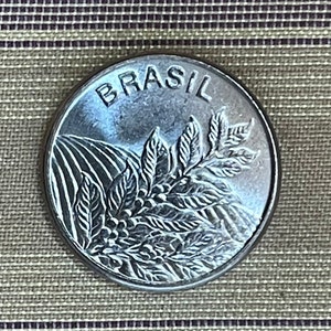 Puede incluir: Una moneda brasileña de plata con la palabra "BRASIL" inscrita alrededor del borde. La moneda presenta una imagen estilizada de una planta de café con hojas y bayas.
