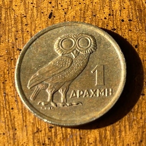 Può includere: Una moneta di bronzo raffigurante un gufo con grandi occhi, il numero 1 e la parola greca "DRACHMI". La moneta ha una forma circolare e un aspetto leggermente usurato, suggerendo che si tratta di una moneta antica.