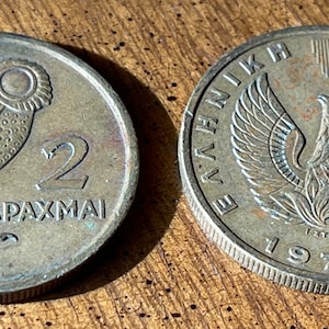 May include: Two vintage Greek 2 Drachmai coins. One coin features an owl and the text "2 DRACHMAI". The other coin has a phoenix design and the text "&Epsilon;&Lambda;&Lambda;&Eta;&Nu;&Iota;&Kappa;&Eta; &Delta;&Eta;&Mu;&Omicron;&Kappa;&Rho;&Alpha;&Tau;&Iota;&Alpha; 1973". Both coins are silver in color.