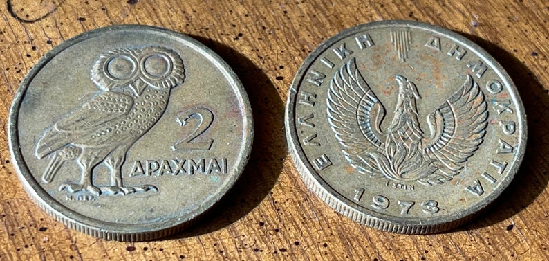 May include: Two vintage Greek 2 Drachmai coins. One coin features an owl and the text "2 DRACHMAI". The other coin has a phoenix design and the text "&Epsilon;&Lambda;&Lambda;&Eta;&Nu;&Iota;&Kappa;&Eta; &Delta;&Eta;&Mu;&Omicron;&Kappa;&Rho;&Alpha;&Tau;&Iota;&Alpha; 1973". Both coins are silver in color.