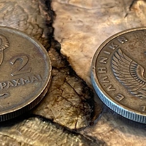 May include: Two vintage Greek drachma coins. One coin features an owl with the number 2 and the word DRACHMAI. The other coin has a phoenix design and the year 1973. Both coins are bronze-colored and show signs of wear.