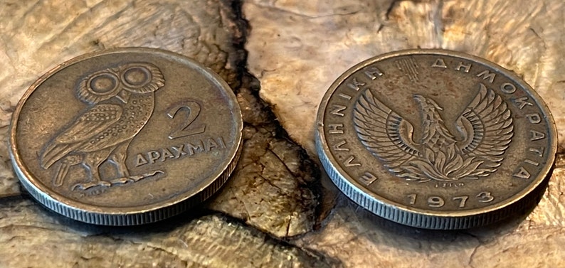 May include: Two vintage Greek drachma coins. One coin features an owl with the number 2 and the word DRACHMAI. The other coin has a phoenix design and the year 1973. Both coins are bronze-colored and show signs of wear.