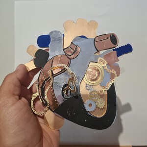 以下が含まれることがあります： 歯車、チェーン、懐中時計が付いた銀と銅色のハート型の彫刻。彫刻は木でできており、スチームパンクの美学があります。