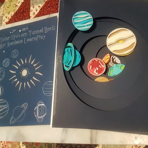 May include: A blue and white paper craft depicting the solar system. The craft features a sun with rays, planets, and a tunnel book design. The title of the craft is "Solar System Tunnel Book by Maniacs LearnPlay".