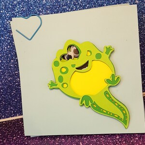 Pode incluir: Um sapo verde e amarelo com olhos saltitantes &eacute; cortado de papel e colado em um cart&atilde;o azul claro. O sapo est&aacute; sorrindo e tem uma cauda longa.