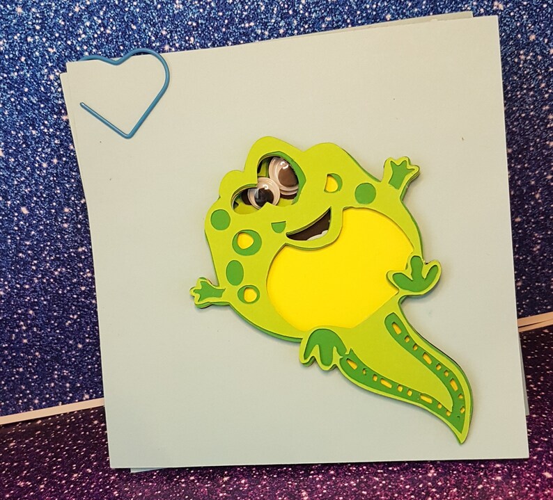 Pode incluir: Um sapo verde e amarelo com olhos saltitantes &eacute; cortado de papel e colado em um cart&atilde;o azul claro. O sapo est&aacute; sorrindo e tem uma cauda longa.