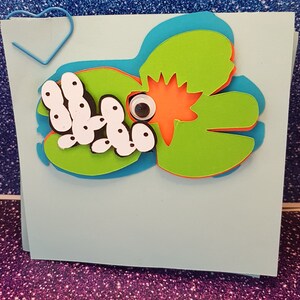 Pode incluir: Um cart&atilde;o azul claro com uma arte de papel verde, laranja e azul de um sapo com um olho saltitante e uma lagarta branca com pontos pretos.