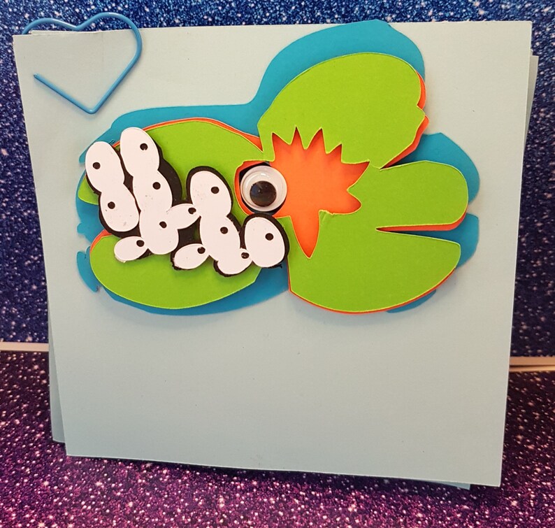 Pode incluir: Um cart&atilde;o azul claro com uma arte de papel verde, laranja e azul de um sapo com um olho saltitante e uma lagarta branca com pontos pretos.