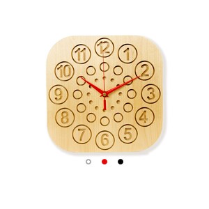 Könnte beinhalten: Quadratische Wanduhr aus hellem Holz. Das Zifferblatt hat eingravierte Zahlen und rote Zeiger. Das Design umfasst unterschiedlich große kreisförmige Aussparungen. Ein dekorativer Artikel für die Inneneinrichtung.