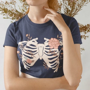 May include: A navy blue t-shirt with a white ribcage design and floral accents. The ribcage is surrounded by pink and white flowers and leaves.