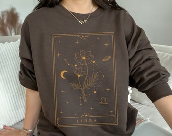 May include: A brown sweatshirt with a gold graphic of a flower and the zodiac symbol for Libra. The graphic is surrounded by stars and a crescent moon. The text "LIBRA" is printed below the graphic.