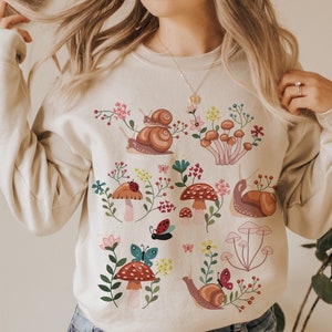 Puede incluir: Una sudadera beige con un dise&ntilde;o colorido de setas y caracoles. El dise&ntilde;o incluye setas rojas y blancas, caracoles marrones, hojas verdes y mariposas de colores.