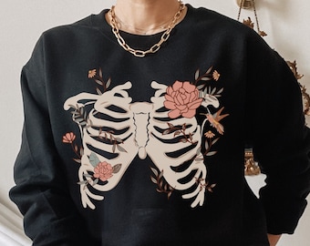 May include: Black sweatshirt with a white ribcage design and floral accents. The ribcage is surrounded by pink flowers, green leaves, and a small hummingbird.