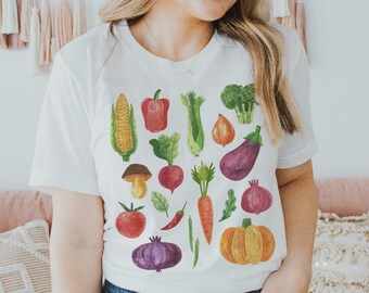 May include: A white t-shirt featuring a colourful watercolour design of various vegetables and fruits. The design includes a red pepper, corn, aubergine, onion, tomato, carrot, pumpkin, and more.