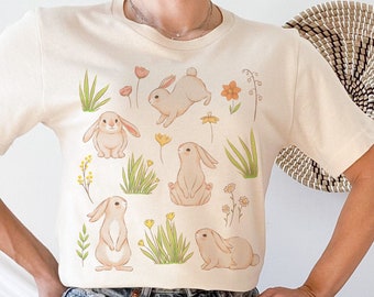 May include: A cream colored t-shirt with a graphic of multiple bunnies and flowers. The shirt is being worn by a person wearing blue jeans.
