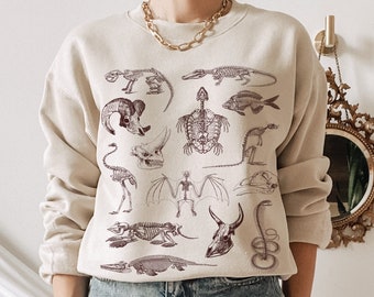 Op de afbeelding: Een beige sweatshirt met een print van verschillende dieren skeletten in bruin. De sweatshirt heeft lange mouwen en een ronde hals.