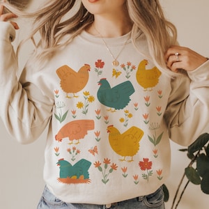 May include: A beige sweatshirt with a colorful pattern of chickens and flowers. The chickens are in various colors, including yellow, orange, blue, and green. The flowers are in shades of pink, red, and yellow.