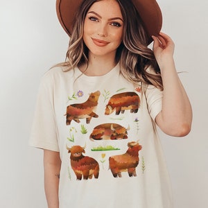 Puede incluir: Una mujer con una camiseta blanca con un dise&ntilde;o de vaca de las tierras altas de color marr&oacute;n, naranja y amarillo. La camiseta tiene cuello redondo y mangas cortas. La mujer lleva un sombrero marr&oacute;n y tiene el pelo largo y casta&ntilde;o.