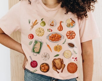 Può includere: Una maglietta rosa con tema del Ringraziamento. La maglietta presenta una varietà di cibi del Ringraziamento, tra cui un tacchino, purè di patate, sugo, salsa di mirtilli rossi e torta di zucca. I cibi sono disposti in modo sparso sulla maglietta.