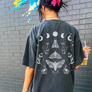Op de afbeelding: Een zwart T-shirt met een witte grafische print van motten, paddenstoelen en manen. De grafiek bevindt zich in het midden op de achterkant van het shirt.