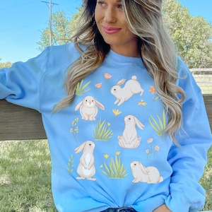May include: A light blue sweatshirt with a graphic print of white bunnies and flowers. The sweatshirt is worn by a person with long blonde hair.