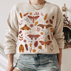 May include: Beige sweatshirt with a colorful graphic of moths, crystals, and foliage. The design features various brown, orange, and pink elements. The sweatshirt has long sleeves and a crew neck. The model is wearing denim shorts.