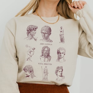 May include: A beige sweatshirt featuring a graphic print of nine different classical sculptures. The text "Ars Longa Vita Brevis" is printed on the sweatshirt.