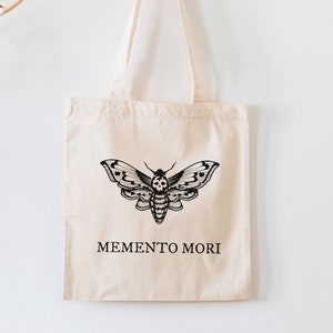May include: A beige tote bag with black straps and a black moth design. The bag has the text "MEMENTO MORI" printed on the front.