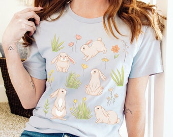 May include: A light blue t-shirt with a pattern of bunnies and flowers. The bunnies are in various poses, including sitting, standing, and hopping. The flowers are in shades of pink, yellow, and orange.