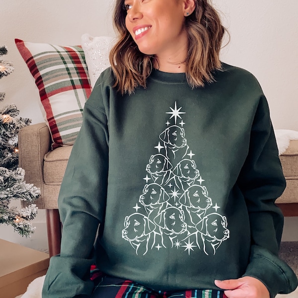 Könnte beinhalten: Ein grünes Sweatshirt mit einer weißen Umrandung eines Weihnachtsbaums aus Hunden. Der Baum hat einen Stern oben und die Hunde schauen alle nach oben. Die Person, die das Sweatshirt trägt, sitzt auf einem Sofa mit einem karierten Kissen dahinter.