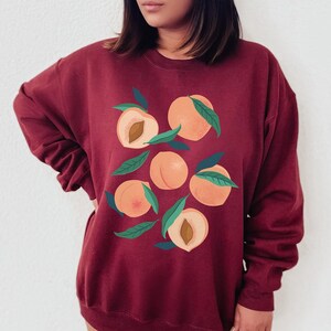 K&ouml;nnte beinhalten: Ein maroonfarbener Pullover mit einem Grafikdruck von Pfirsichen. Die Pfirsiche sind in einer Traube angeordnet und haben gr&uuml;ne Bl&auml;tter.