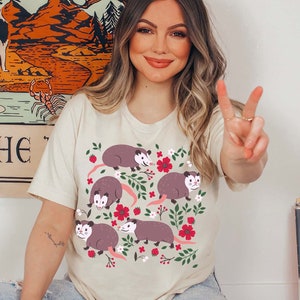 May include: A beige t-shirt with a graphic print of multiple opossums with pink flowers and green leaves. The opossums are brown with white accents.